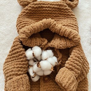 May include: A brown, hooded baby coat with bear ears, made of a soft, textured material. The coat has a button closure and is adorned with a sprig of cotton. The text "@Gu Suknitting" is visible on the bottom right.