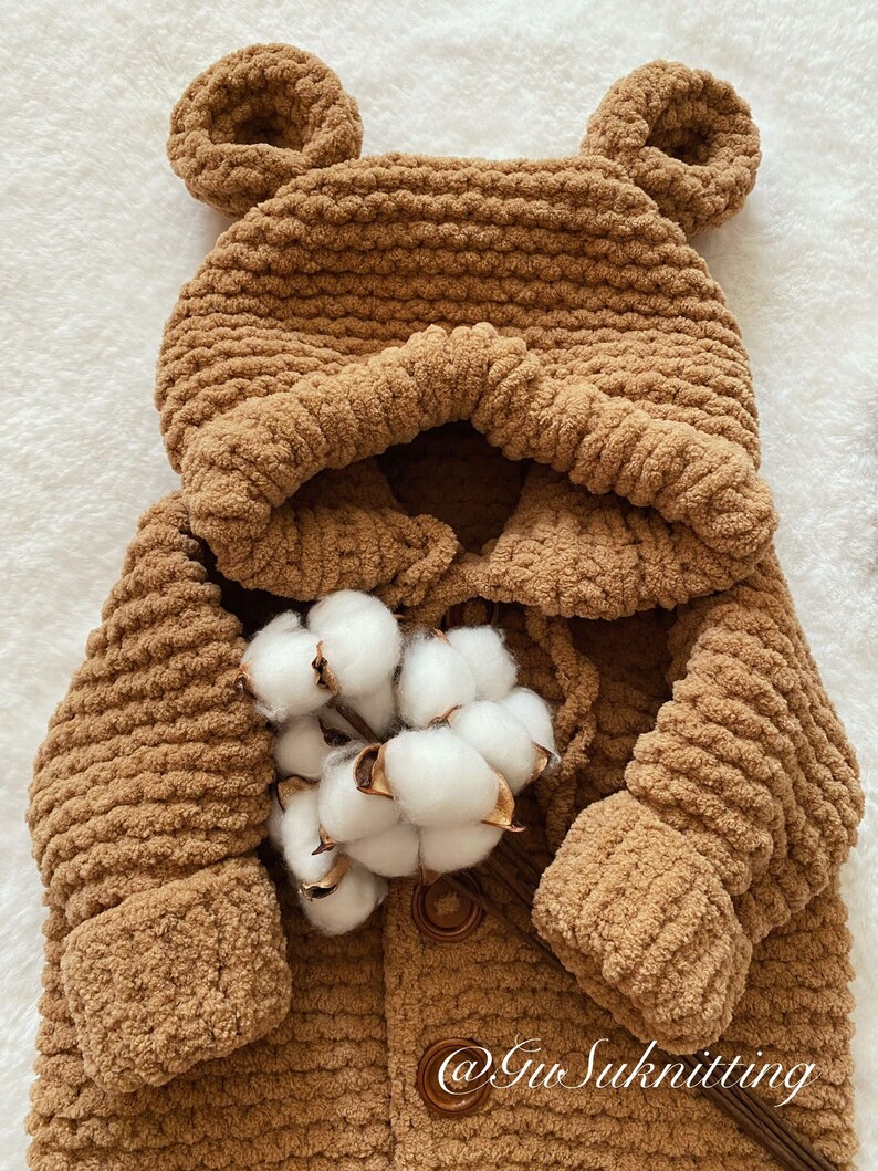 May include: A brown, hooded baby coat with bear ears, made of a soft, textured material. The coat has a button closure and is adorned with a sprig of cotton. The text "@Gu Suknitting" is visible on the bottom right.