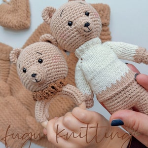 May include: Crocheted teddy bear rattle and stuffed bear in beige and white. The rattle has a wooden ring and a brown collar. The stuffed bear wears a white sweater with beige accents. Other matching crocheted items are in the background.