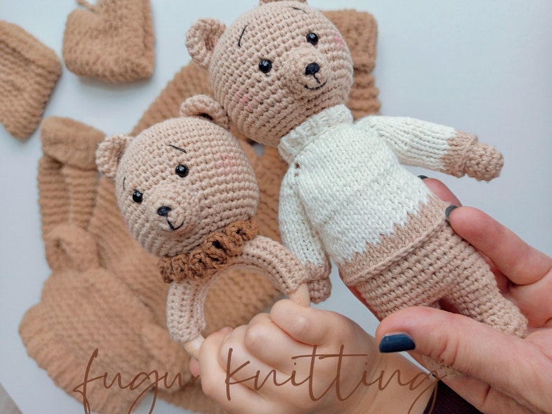 May include: Crocheted teddy bear rattle and stuffed bear in beige and white. The rattle has a wooden ring and a brown collar. The stuffed bear wears a white sweater with beige accents. Other matching crocheted items are in the background.