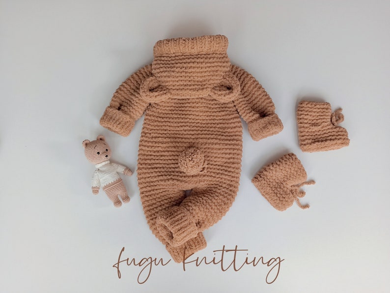 May include: A light brown, knitted baby outfit set, including a jumpsuit with bear ears, booties, and a small teddy bear toy. The jumpsuit has long sleeves and a turtleneck collar. The booties have a tie closure. The teddy bear is wearing a white shirt.