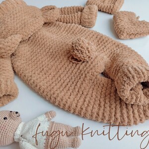 May include: A light brown, plush knitted baby outfit set, including a jumpsuit, hat, and booties. The set is made of soft, textured yarn. A small, crocheted teddy bear is also present. The text "fugu knitting" is visible.