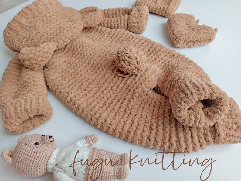 May include: A light brown, plush knitted baby outfit set, including a jumpsuit, hat, and booties. The set is made of soft, textured yarn. A small, crocheted teddy bear is also present. The text "fugu knitting" is visible.