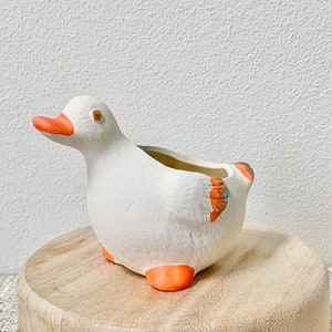 May include: A white ceramic duck planter with orange beak, feet, and wing accents. The duck-shaped planter has a hollowed-out top for planting small plants or succulents. The planter is sitting on a round wooden base.