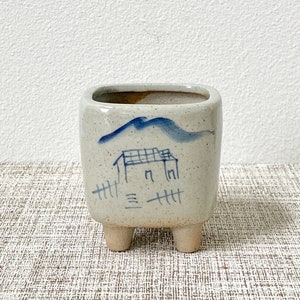 Peut inclure: Un petit pot en céramique blanc avec un motif de montagne et de maison bleu et blanc. Le pot a trois petits pieds et est parfait pour une petite plante grasse ou un cactus.