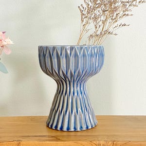May include: A blue ceramic vase with a unique hourglass shape. The vase features a textured design with vertical and geometric patterns. Dried flowers are placed inside the vase, adding a natural element to the decor. The vase is placed on a wooden surface.
