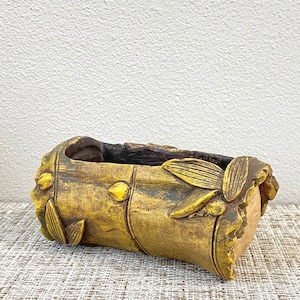 May include: A handcrafted, rectangular planter with a bamboo-like design. The planter is primarily yellow-brown with darker brown accents and leaf-shaped embellishments. It is suitable for small plants or succulents.