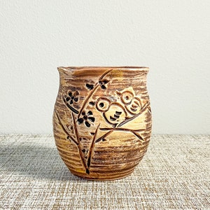 May include: A brown ceramic vase with a textured surface. The vase features a carved design of birds perched on a branch with flowers. The color palette includes shades of brown, tan, and black.