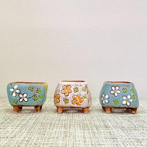 May include: Three small, ceramic planters with a square shape and short legs. The planters are decorated with floral patterns in white, orange, and green on a blue and off-white background. The planters are arranged on a textured surface.