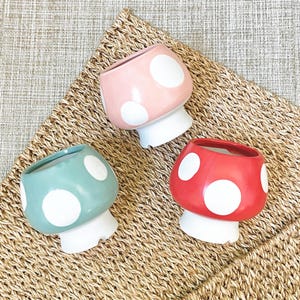 May include: Three ceramic mushroom-shaped planters in pastel colors: pink, teal, and red. Each planter has white polka dots and a white base. The planters are arranged on a woven, tan surface.