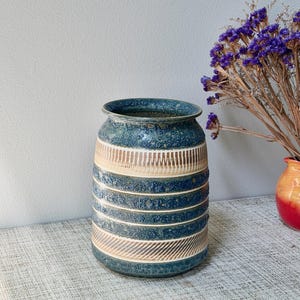 May include: A cylindrical ceramic vase featuring a textured blue glaze with horizontal bands of cream-colored incised lines and rough, dark blue speckled sections. The vase has a wide, slightly flared rim and a flat base, displaying a pattern of alternating smooth and textured stripes.