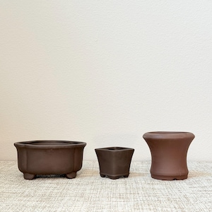 May include: Three unglazed brown ceramic pots of varying shapes. The left pot is rectangular with rounded corners and four small feet. The center pot is a small square with four feet. The right pot is cylindrical with a flared rim.