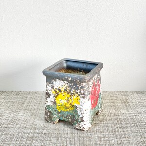 May include: Square ceramic planter with a distressed white finish and painted floral designs in yellow, red, and green. The planter has four small feet.