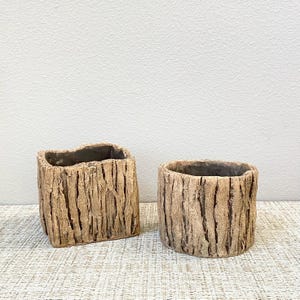 May include: Two decorative planters with a textured, bark-like appearance. One is square, the other is round. Both are light brown with dark brown vertical lines, creating a natural, rustic aesthetic. The planters are suitable for small plants or succulents.