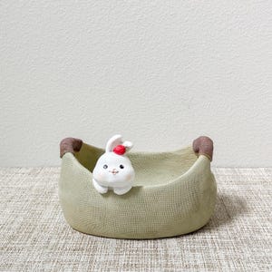 May include: A small, green ceramic planter shaped like a bag with brown handles. A white ceramic bunny with a red berry on its head is peeking out of the planter.