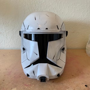 May include: A white and black helmet with a visor, possibly a costume item. The helmet has a rounded top and angular features, with black accents and simulated wear marks. The visor is black, and the helmet appears to be made of a hard material.
