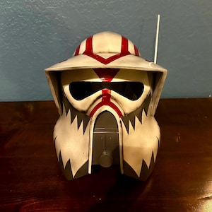 Puede incluir: Un casco blanco y gris con rayas rojas y una visera negra. El casco tiene una parte superior puntiaguda y un ala ancha. Es un casco de soldado clon de Star Wars.