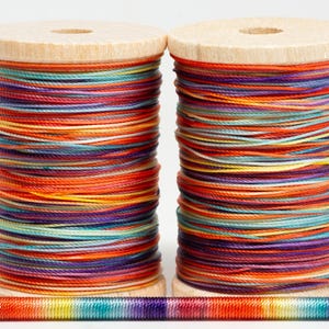 May include: Two wooden spools of colorful thread, featuring a vibrant array of orange, red, purple, and teal hues. The thread is tightly wound, creating a textured appearance. A small section of the thread is unraveled at the bottom, showcasing the color variations.