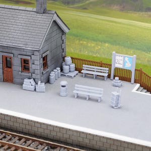 May include: A detailed miniature model railway station scene. It features a grey stone building, benches, barrels, and a signpost. The backdrop shows a green field and a blue sky, creating a realistic diorama.