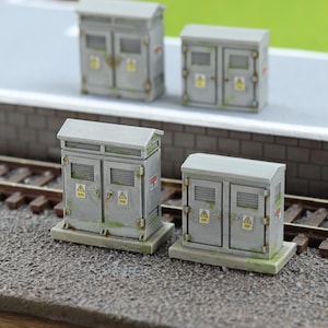 May include: Miniature grey electrical cabinets with hinged doors, yellow warning symbols, and a weathered appearance. The cabinets are set on a concrete base, with a roof and are placed near a railway track.