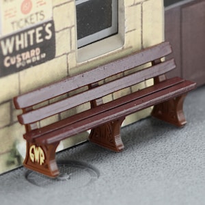 May include: A brown wooden bench with a GWR logo on the side. The bench is in front of a building with a sign that says "Whites Custard Powder".