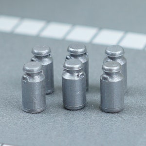 May include: Six miniature silver milk churns with rounded lids and handles. The small jugs are arranged on a grey surface, suggesting a collection or decorative item. The overall aesthetic is simple and industrial.