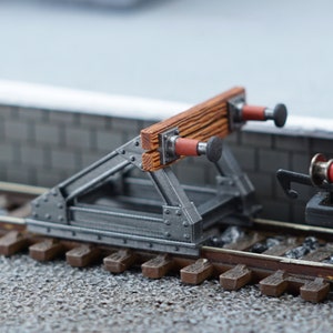 May include: A model railway buffer stop, with a wooden beam and a metal frame. The buffer stop is painted in brown, red, and grey. It is positioned on a model railway track. The scene includes a section of a model train.