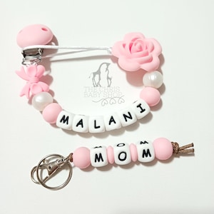May include: A pacifier clip featuring a pink silicone rose, a pink bow, white and pink beads, and white letter beads spelling MALANI, paired with a matching keychain featuring white letter beads spelling MOM and a silver metal keyring.