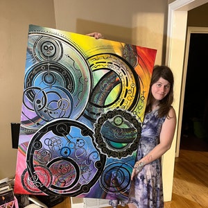 May include: A large abstract painting with a colorful, geometric design featuring overlapping circles and swirls in shades of blue, green, yellow, pink, and black. The painting is held by a person in front of a wooden floor.
