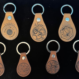 May include: A collection of seven brown leather keychains with silver key rings. Each keychain features a unique black graphic design and text, including "T.A.R.D.I.S", "Always", and "Hello Sweetie". The keychains are attached with silver rivets.