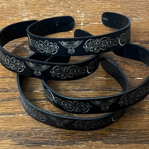 May include: Four black metal cuff bracelets with engraved silver designs. Each bracelet features a repeating pattern of stylized birds and Celtic knotwork. The bracelets are stacked on a wooden surface, showcasing their open cuff design.