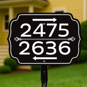 May include: Black address sign with white numbers 2475 and 2636, with arrows indicating direction. The sign is mounted on a black post and set in a green lawn. The background features a yellow house.