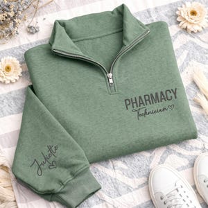 May include: A sage green quarter-zip sweatshirt featuring a silver zipper and ribbed cuffs. The chest displays the black text "PHARMACY Technician" with a small heart icon, while the sleeve is embroidered with the name "Juliette" and a heart.