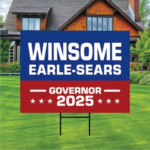 May include: A blue and red political campaign sign with the words "WINSOME EARLE-SEARS" in white. Below, it reads "GOVERNOR 2025" with stars. The sign is in a garden with a house in the background.