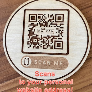 May include: A wooden coaster with a black and white QR code that says "Cafe Balkan - Authentic Balkan Kitchen" and the text "Scan Me" below the QR code. The text "Scans to your personal website address!" is written in red below the coaster.