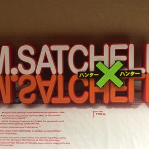 May include: A red and orange sign with the text "M. SATCHELL" and "W241CHET" in white letters. The sign also has a green "X" with the text "ハンター" in yellow on either side.