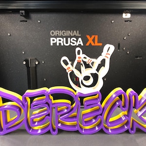 May include: A 3D printed name sign that says "Derek" in purple and yellow. The sign is sitting in front of a black 3D printer with the words "ORIGINAL PRUSA XL" printed on the front.