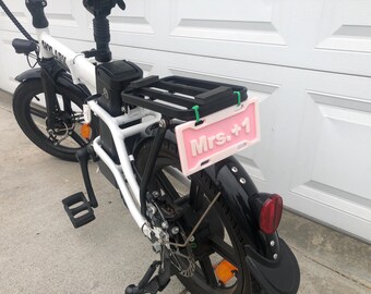May include: White electric bicycle with a black rear rack and a pink license plate that says "Mrs. +1".