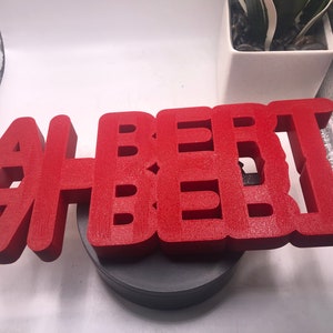 May include: A red 3D printed sign that reads "ALBERT" in all capital letters. The sign is sitting on a black circular base.