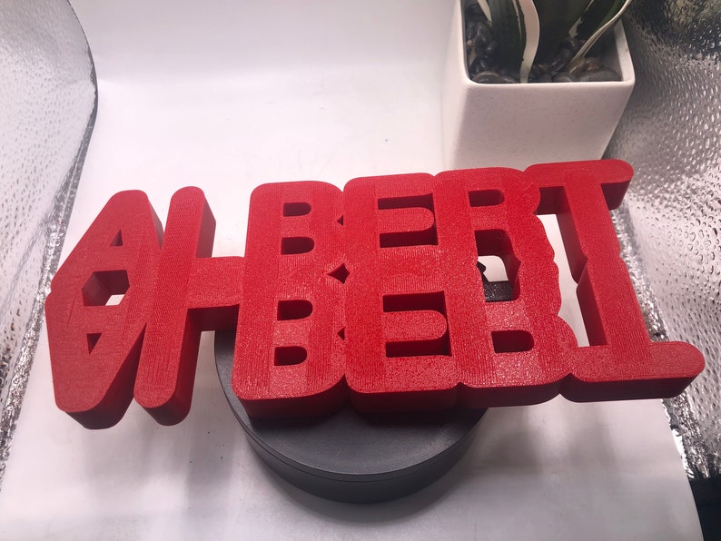 May include: A red 3D printed sign that reads "ALBERT" in all capital letters. The sign is sitting on a black circular base.
