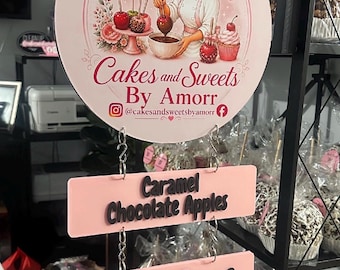 May include: A round pink sign featuring an illustration of a baker dipping an apple, with the text Cakes and Sweets By Amorr and social media handles. Below are three rectangular pink signs with black text: Caramel Chocolate Apples, Cake Pops, and And more...