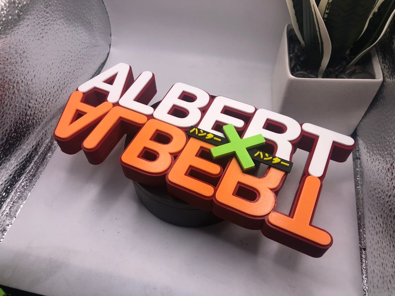 May include: A 3D printed sign with the words "ALBERT VIBERT" in white and orange letters. The letters are layered on top of each other, creating a dimensional effect. The sign also features a green "X" with the Japanese word "ハッダー" written in yellow on either side of the "X".