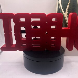 May include: A red, 3D, wooden sign that reads "BE THE" with an arrow pointing to the right. The sign is on a black, round base.