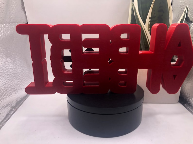 May include: A red, 3D, wooden sign that reads "BE THE" with an arrow pointing to the right. The sign is on a black, round base.