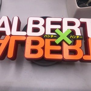 May include: A 3D printed sign with the word "ALBERT" in white letters on top of the word "ALBERT" in orange letters with a green "X" in the middle. The word "ハッカー" is written in black on either side of the "X".