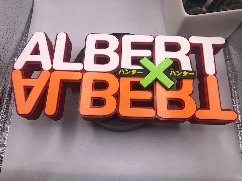 May include: A 3D printed sign with the word "ALBERT" in white letters on top of the word "ALBERT" in orange letters with a green "X" in the middle. The word "ハッカー" is written in black on either side of the "X".
