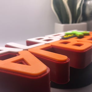 May include: A close-up of a 3D printed name sign that says "ALBERT" in orange and white letters with a red base.