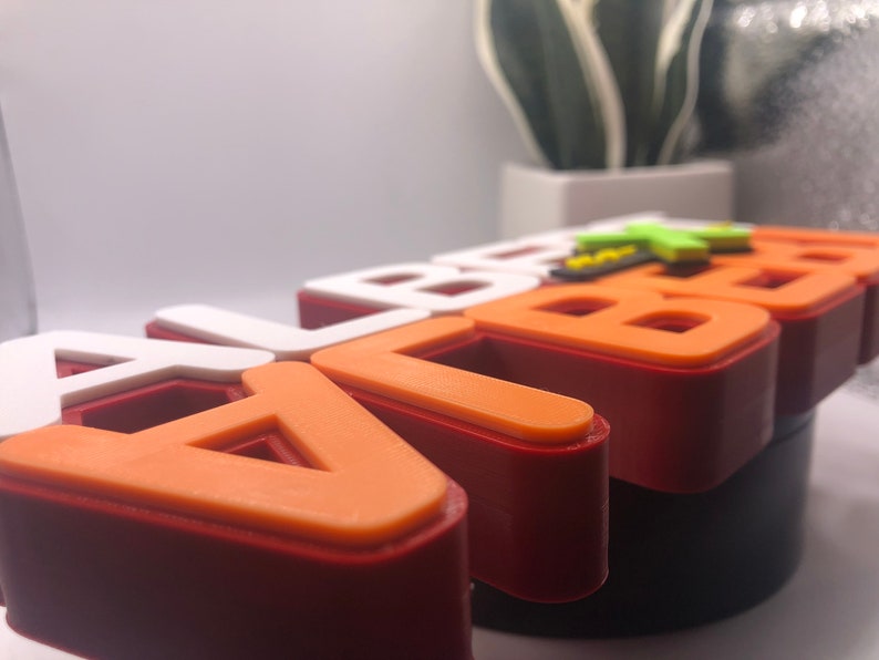 May include: A close-up of a 3D printed name sign that says "ALBERT" in orange and white letters with a red base.