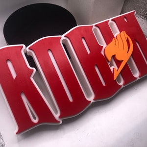 May include: A 3D printed red and white sign with the word "FAIRY TAIL" in a stylized font. A yellow and orange fairy wing symbol is on the right side of the sign.