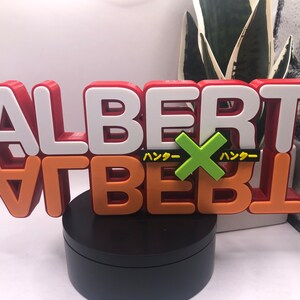 May include: A 3D printed red, white, and orange sign with the word "ALBERT" in white letters on top of the word "ALBERT" in orange letters. The sign has a green "X" with the Japanese word "ハッダー" on either side of the "X". The sign is on a black circular base.